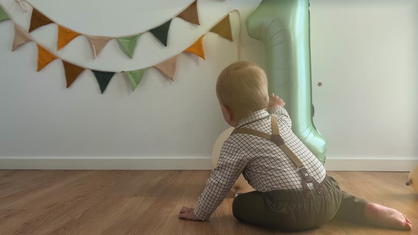 Cute baby boy celebrates 1 year birthday. Toddler plays among balloons and sage green inflatable number one. Child in beautiful costume and festive decor. 