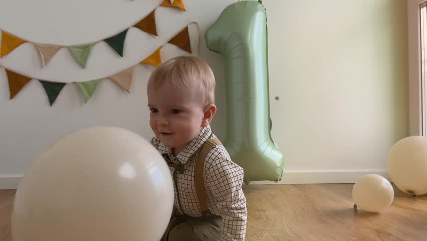 Cute baby boy celebrates one year birthday. Toddler plays in sunny room among balloons and sage green inflatable number one. Child in beautiful green suit. 