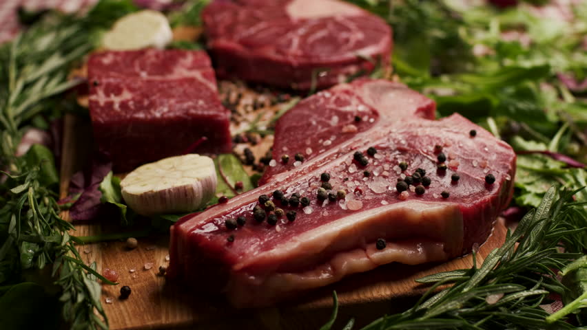 Raw T-Bone Beef Steak Being Salted with Greenery Around