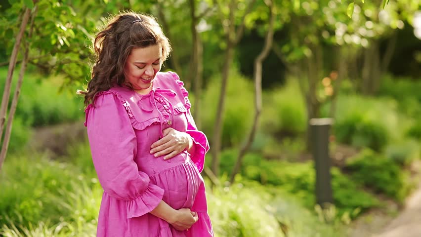 Beautiful and happy pregnant young woman in pink dress in the park. Long-awaited and healthy pregnancy. clothes and cosmetics for expectant mothers.