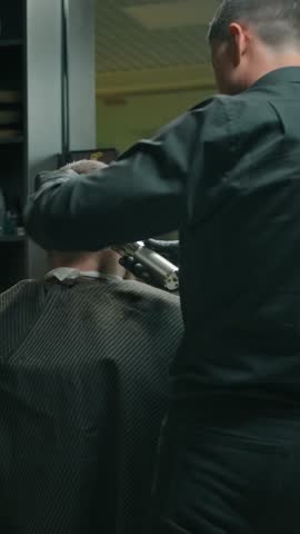 Middle shot from the back of a client haircut by a hairdresser. Work in a modern barbershop, client and barber. Two masters in front of the mirrors trim their customers with a trimmer. Vertical shot.