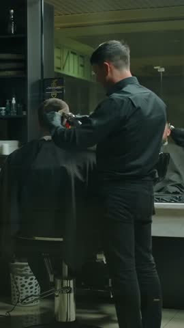 Two clients and two hairdressers haircut in a dark barbershop room. Dolly Camera overall plan, work of two professional barbers and pink-haired client and also a mid-aged client. 4 People Vertical