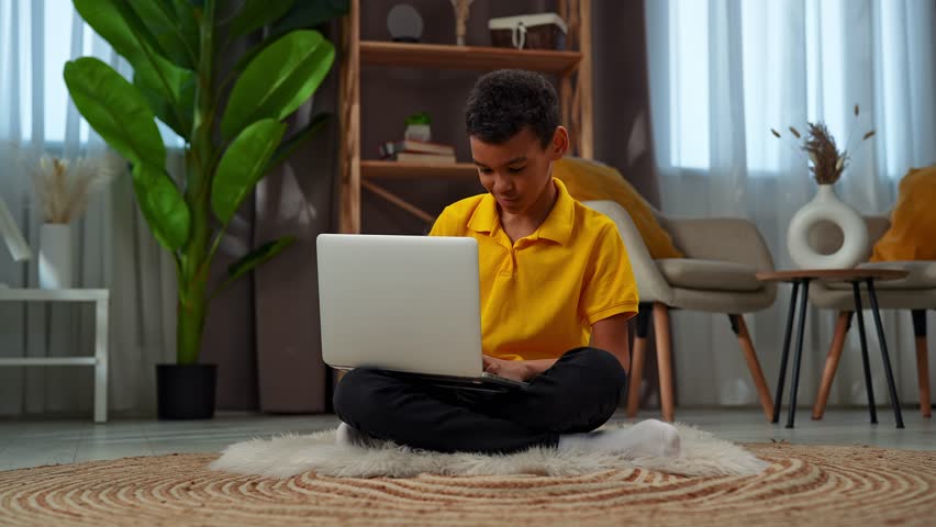 Daily lifestyle concept. Young african american child boy spending free time at home, kid sits on the floor in the living room and texting online on laptop, positive smiling expression.