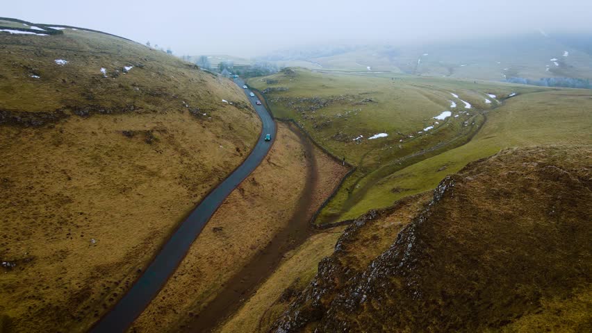 Cars driving along curving road through grassy hills. Vehicles cruising on scenic route in Peak District, United Kingdom. Green and red cars traveling on smooth asphalt path surrounded by rugged