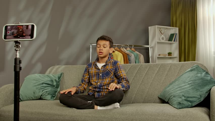 Daily lifestyle concept. Young african american child boy spending time at home, kid blogger sits on sofa records video on smartphone, talking at camera asking for likes and subscribe.