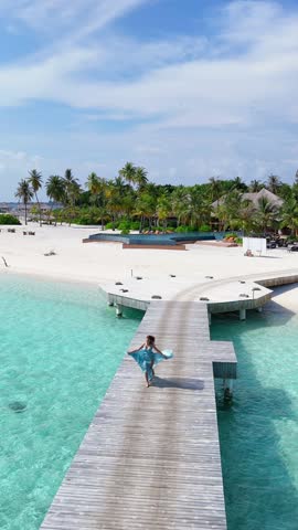 Happy woman in flowing airy dress runs along wooden pier over clear turquoise water toward tropical island, joyful escape in Maldives, summer vacation, traveling, relax