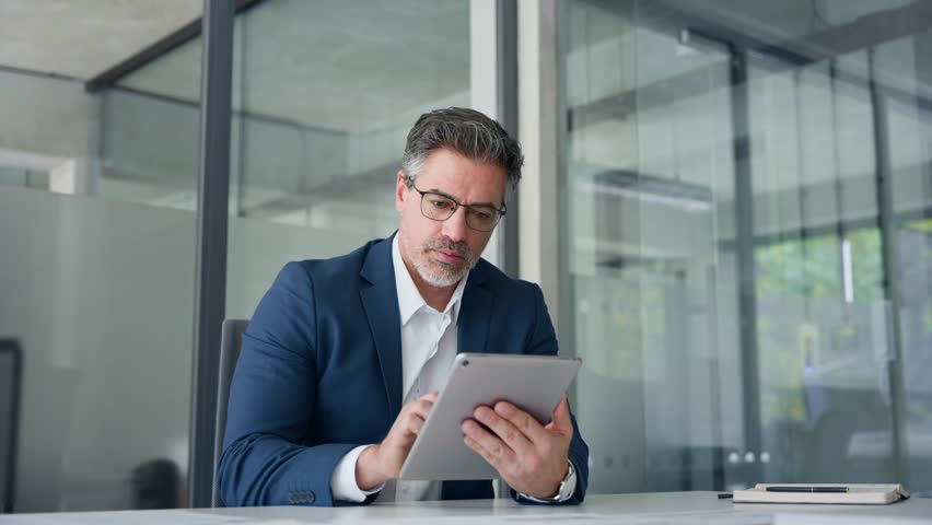 Positive senior businessman entrepreneur sitting at desk working on financial sales project, looking smiling at screen. Mature 50s Latin business man ceo manager using tablet pad computer in office