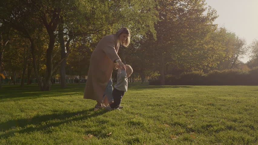 Nurturing mother gently guiding barefoot toddler while taking first independent steps across sunlit green lawn, supporting child's walking milestone with loving care