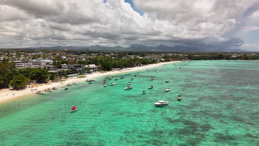 Aerial footage of a popular Mauritius beach town with boats in turquoise lagoon under cloudy sky.Cloudy day over Mauritian bay: Aerial perspective of numerous boats near beach town and mountains.