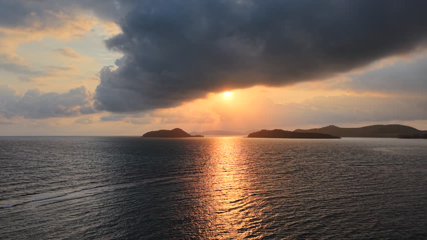Sunbeams breaking through dramatic cloud over sea casting glowing path across surface as waves ripple gently toward island, warm golden light reflecting movement in tropical evening seascape