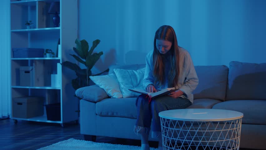 Young Woman Comfortably Sitting on Sofa, Quietly Reading the Holy Bible in Soft Evening Light. Peaceful and Calm Atmosphere for Scripture Study, Spiritual Reflection, Faith, and Personal Devotion.