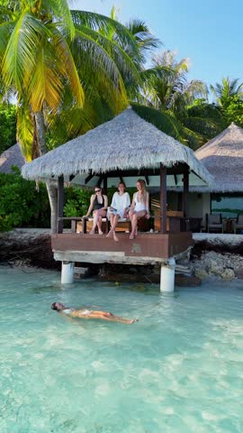 Three women relax on a wooden terrace above turquoise water while another swims nearby, tropical resort in maldives, peaceful vacation, summer retreat, relaxation
