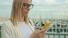 Professional woman in white blazer standing near marina, gripping smartphone with yellow case, scanning digital content during workday break.Business, technology, blogging, communication concept - Powered by Shutterstock - Get 15% off with code: PIKWIZARD15