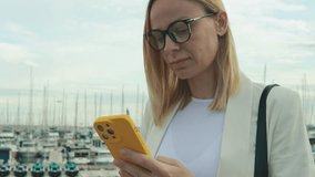 Professional businesswoman in white blazer working on yellow-cased smartphone near marina, checking emails and browsing digital content.Business, technology, blogging, communication concept - Powered by Shutterstock - Get 15% off with code: PIKWIZARD15