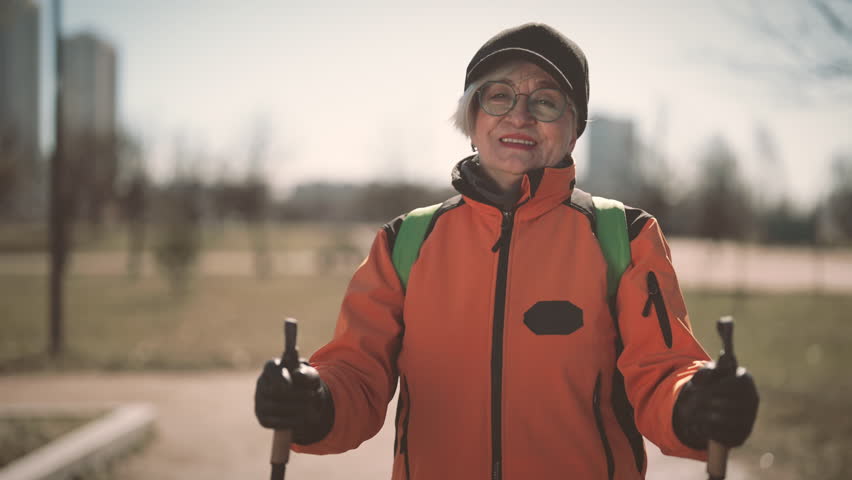 Better aging and sport for retirees, old woman practicing Nordic walk, portrait. Medium shot with charming smiling lady holding sticks for sportive walk, save good body shape in old age, slow motion