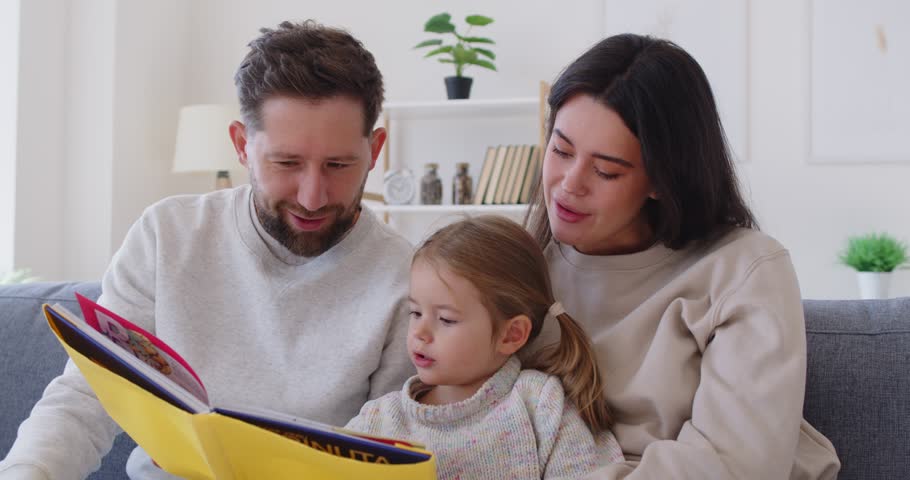 Happy family mother, father and little girl sitting close on sofa and reading interesting book together. Cheerful loving parents spending leisure time with their daughter, enjoying fairytale story.