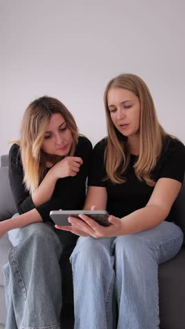 Vertical video. Two adult  women are sitting close to each other on a sofa, focused on their smartphones. Discussing social media.