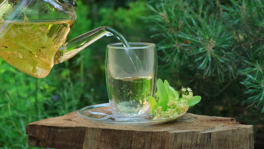 Linden Tea Pouring from Teapot into Glass Cup. Tea ceremony outdoors in summer or spring day. Refreshing Linden Herbal Tea in a Glass Cup