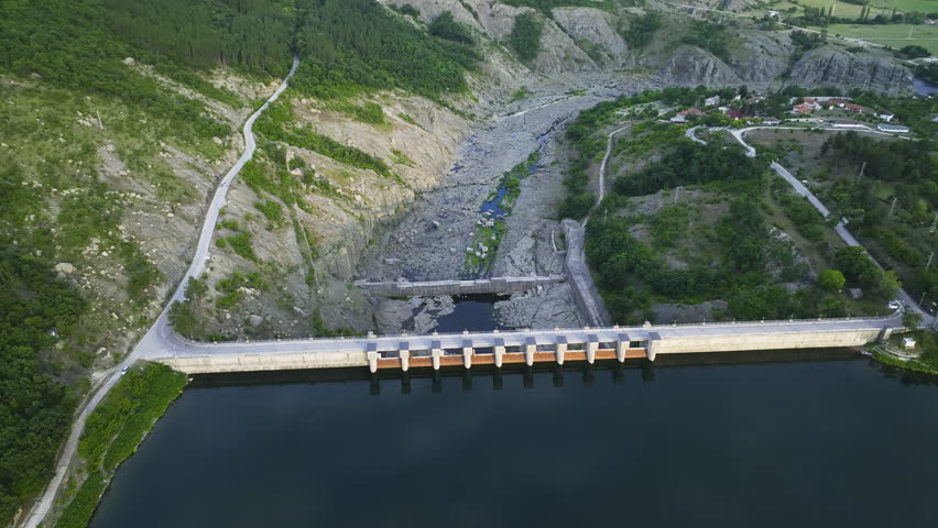 Large hydroelectric dam spanning a calm river with rocky mountainous terrain around it, including roads, greenery, and a visible reservoir in the foreground.