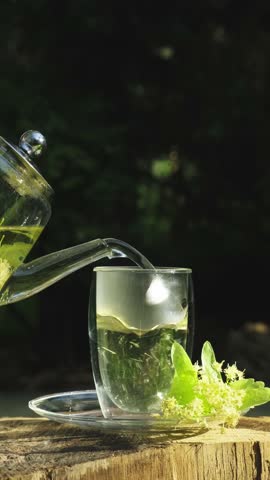 Pouring Linden Tea into Glass Cup Outdoors. Relaxing Linden Herbal Tea. Glass Cup and Teapot in Green Nature. Outdoor Summer Beverage Experience Vertical video