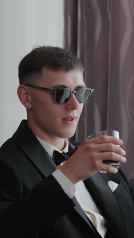 Stylish man in a black suit and sunglasses, drinking in a relaxed indoor setting . Vertical video
