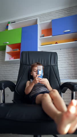 Relaxed Caucasian baby boy sits in a big black leather chair. Kid holds a slider phone and pretends talking on the phone. Vertical video.