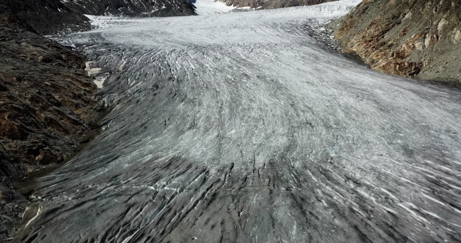 Drone glides low over the glacier’s cracked ice surface, revealing alpine peaks in the distance and dramatic icy textures.