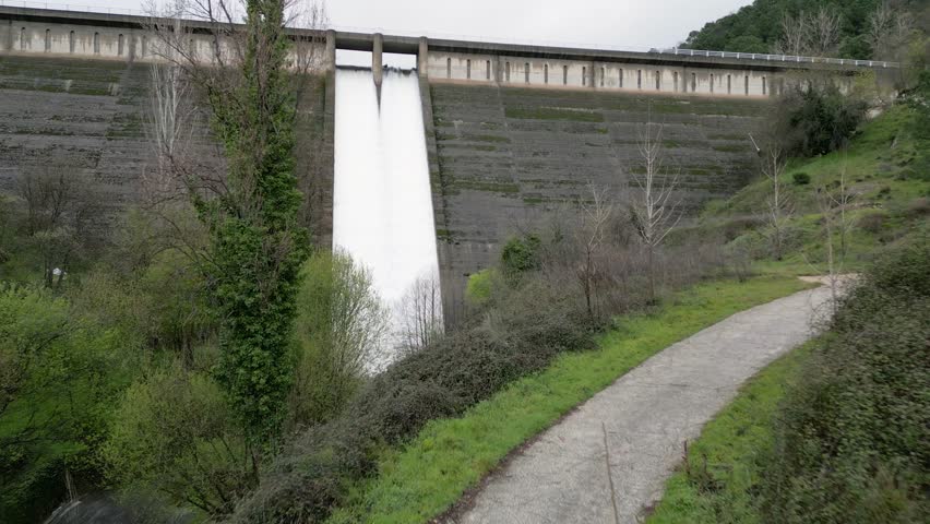 Excellent drone footage of a mountain dam. We see the spectacular waterfall formed by the dam's overflow, which is white and very long due to its height. The green riverbank appears.
