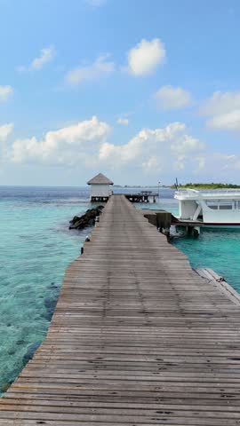Wooden pier stretches over rocks and turquoise sea with boats docked nearby, peaceful marine view in maldives, coastal summer travel, resort escape. 