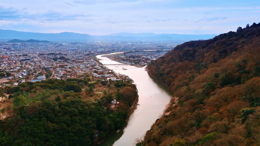 Flight above the narrow Katsura river in Kyoto, Japan. Vast cityscape surrounded by mountains at backdrop. Aerial view.