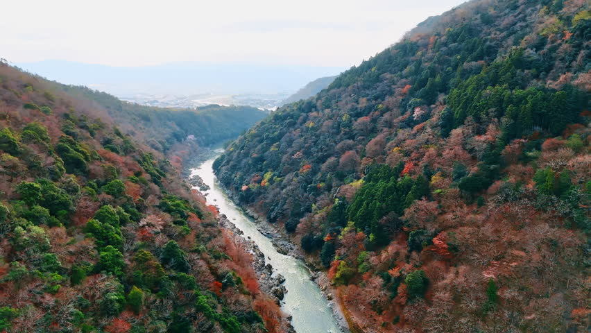 Tranquil autumn in the mountains of Kyoto, Japan. Drone footage above the Katsura river. Cityscape at backdrop.
