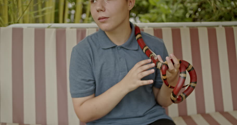 A teenage girl sits on a bench outside, playing with her pet snake.