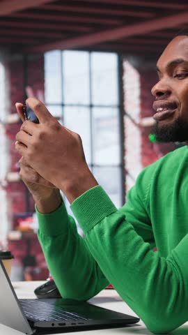 Vertical Video Man at home using smartphone to compose social media posts, producing content as a freelancer. African american guy monitoring profile engagement using mobile phone. Camera B.
