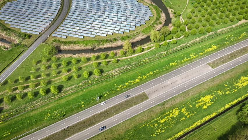 Aerial footage of a ring road near Almere in the Netherlands, showing the roadway with traffic, solar panels, canal and surrounding green areas with trees and yellow flowers in spring.