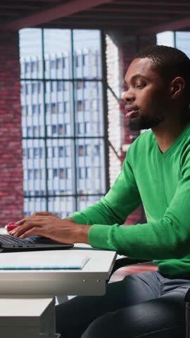 Vertical Video African american focused guy typing on his laptop at home office to solve daily tasks, doing remote freelance work online. Young adult surfing the web multitasking in loft apartment