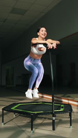 Energetic young fitness girl in white top and blue iridescent leggings doing exercises jumping on mobile mini trampoline. Jumping workout. High intensity interval cardio training, women's health
