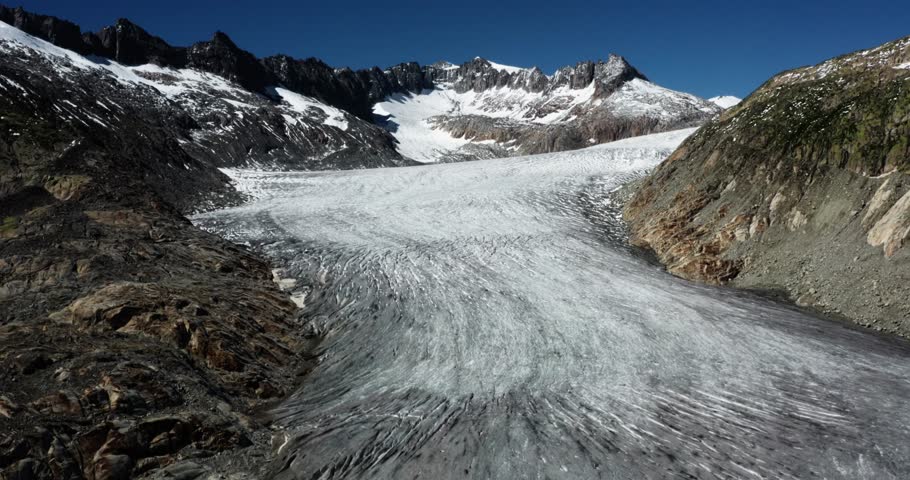 Drone footage showing a wide glacier scene surrounded by majestic Swiss Alps and a vibrant blue sky.