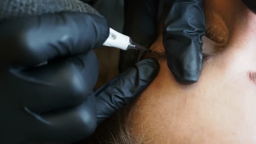 Tightly framed on the moment of contact, this extreme close-up shows the microblading pen etching a precise stroke into the client’s brow as gloved fingers anchor the skin gently, slow motion.