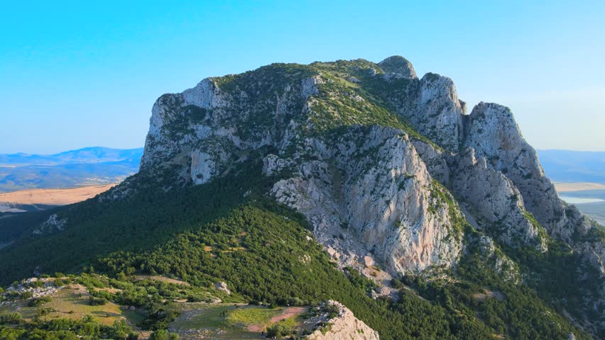 A stunning panoramic view of a rugged, towering rock formation dominating the serene Tunisian landscape, surrounded by lush greenery and expansive plains under a clear blue sky.