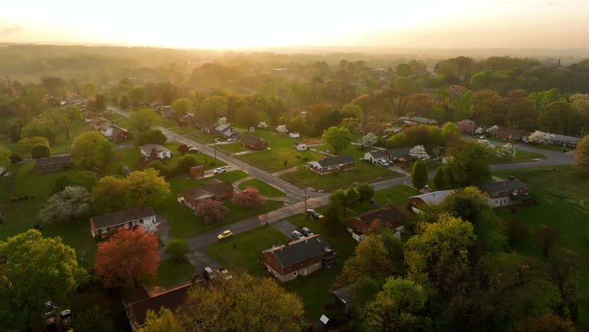 Quiet American suburb neighborhood with one family houses during golden sunset. Aerial top down shot. Peaceful housing area with glowing sunlight and fog. Spring day with colored trees. Virginia, USA.