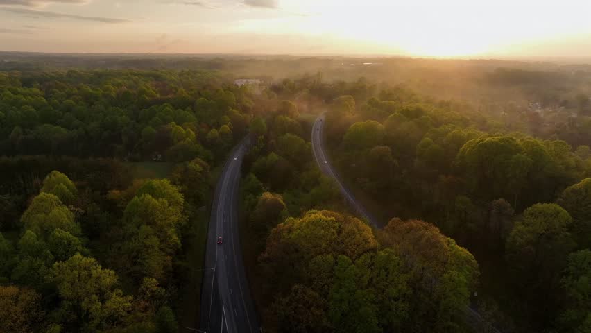 Slow drone flight over american Highway between green trees at golden sunset. Aerial wide shot. Cars on roads between forest and swaying fog and mist. Quiet rural landscape in USA.