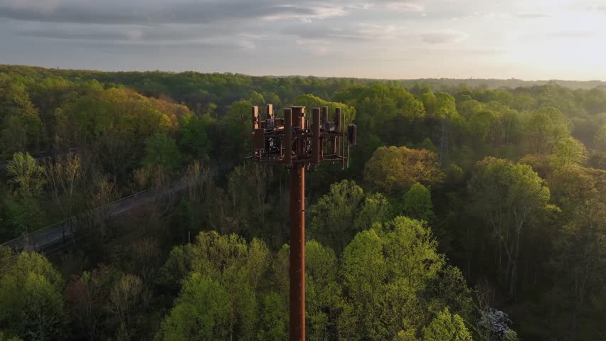Aerial orbit shot of transmission tower in forest landscape of America. Driving cars on rural road during golden Sunset. Green colored trees in spring season.Fog and mist on mystic evening in USA.