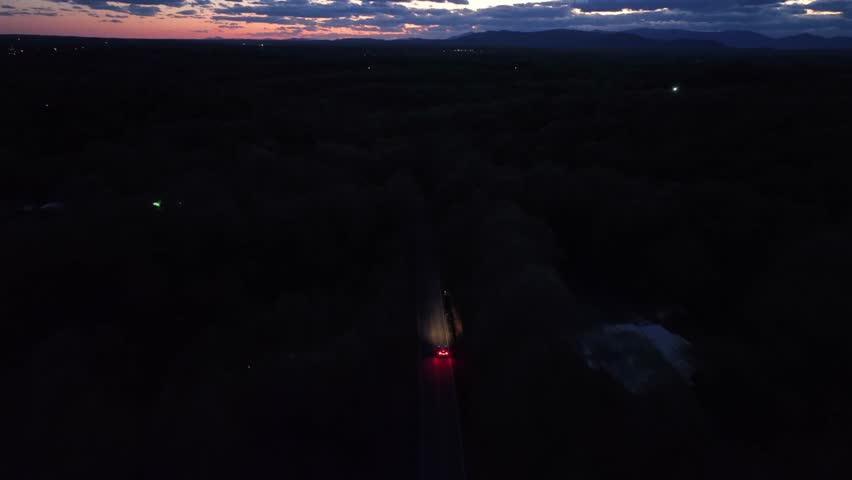 Aerial following shot of car on dark street between dense dark forest trees at night. Sunset time at sky. Drone tracking shot. Dark housing area of america.
