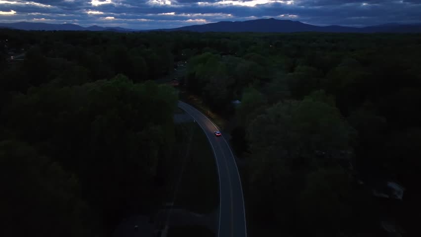 Car with headlights on empty intersection road between forest tress in suburb of American town. Dusk scene with clouds after sunset. Aerial tracking wide shot.