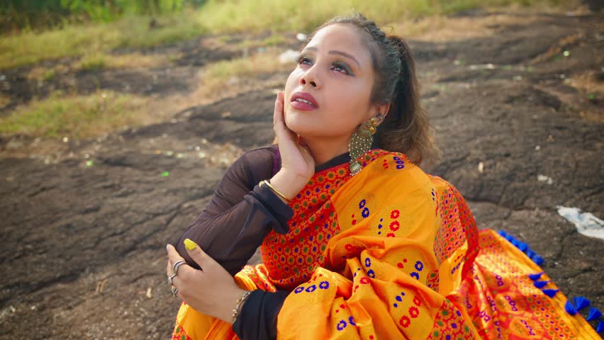 Pensive lady rests on jagged boulder gazing upward, Girl wear ethnic dress remains still with sorrow within, Emotional soul weeps quietly lost in inner storm, Memory rush brings tears as she sit alone