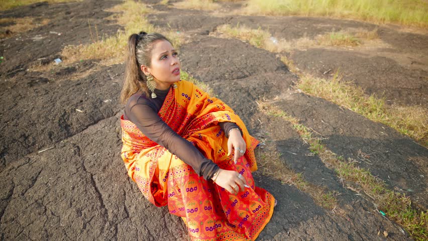 Drone shot woman hums softly wearing sari outdoors, Aerial view singer sits on rock sings with hand motion, Artist in ethnic attire croons holding cigarette, Girl mouthing lyrics by open meadow, Rural