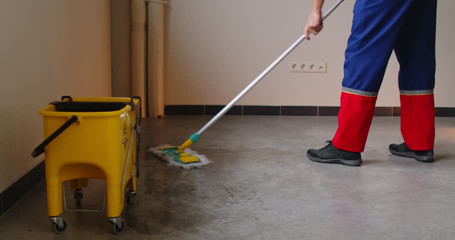 Cropped view of person in work uniform cleaning wet floor with mop in empty room with yellow mop bucket on wheels nearby. Professional janitor performing routine service and floor sanitation work.