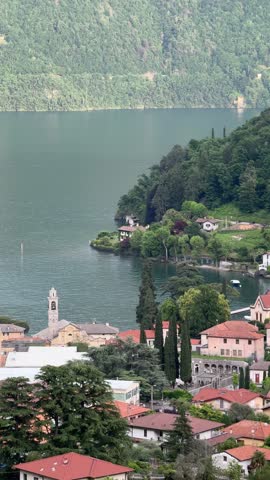 Panoramic view of Lenno town on Lake Como, Lombardy, Italy. Colorful houses, church tower, lush green hills, and calm lake waters on a sunny summer day.