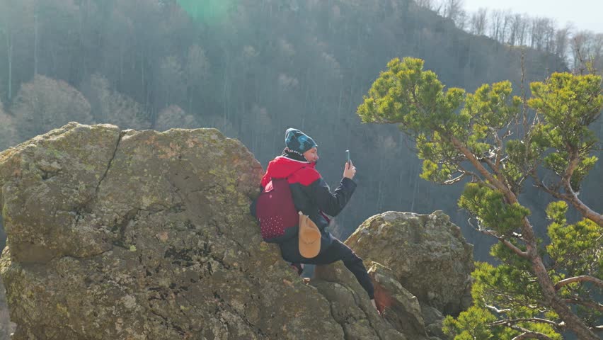 A girl in a bright red jacket sits on a rock ledge and captures the stunning scenery on her phone. Nearby are the branches of a green pine tree. Tourist in the rocky mountains during a hiking trip