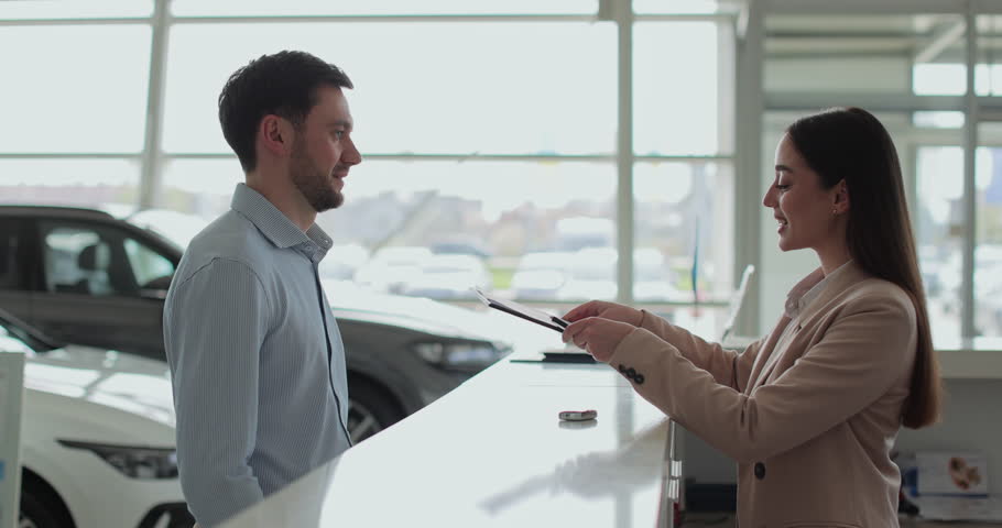 Handsome man buying a new car and signing the contract, car in the background. Saleswoman gives a car keys to buyer. Concept of car shopping, buying, and dealership.